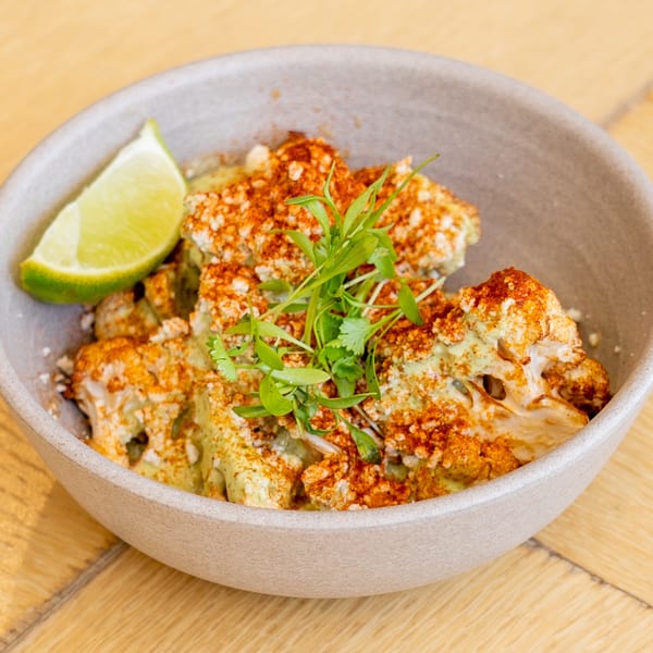 Roasted cauliflower with green sauce, red spice, fresh herbs, and a lime wedge in a bowl.