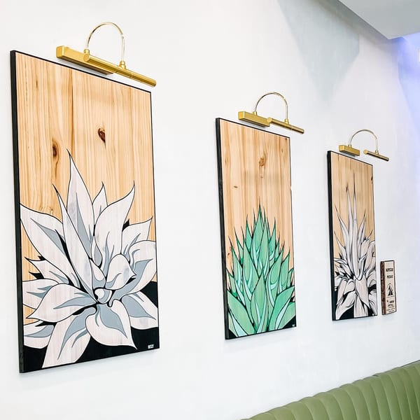 Three agave plant paintings on wood panels with brass lights above, over a green booth.