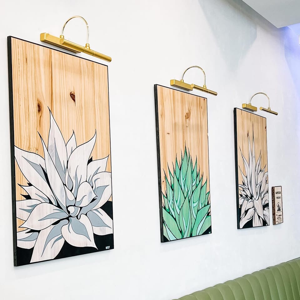 Three agave plant paintings on wood panels with brass lights above, over a green booth.