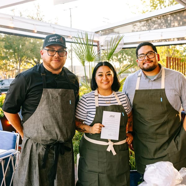 Three smiling El Barrio staff members, two men and one woman, pose outdoors on a sunny day.